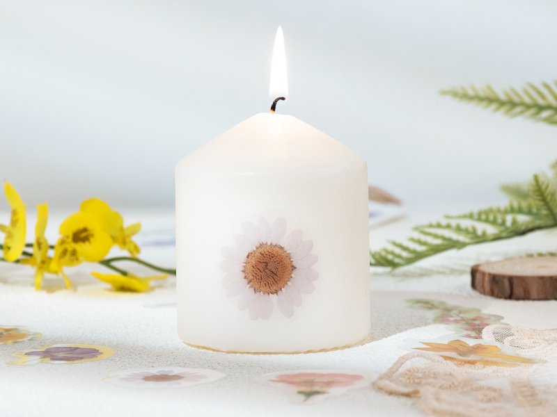 White pillar candle with a daisy sticker, surrounded by floral stickers and greenery, with a lit flame.