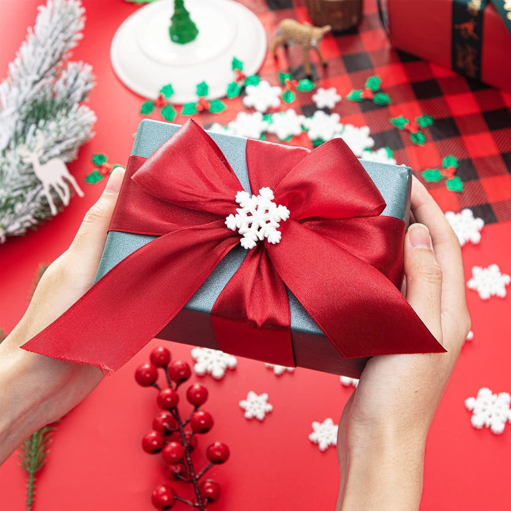 Hands holding a teal gift box with a large red satin bow and a white snowflake ornament, surrounded by scattered snowflakes, holly berries, and a sprig of pine on a red background.