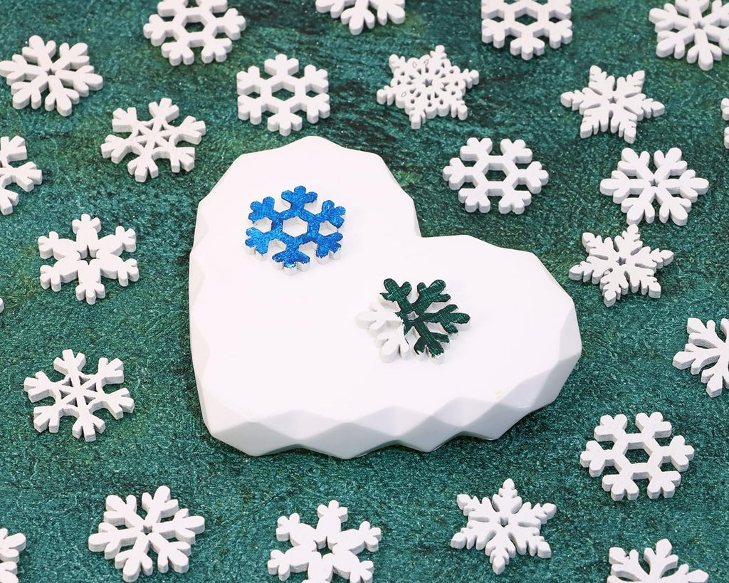 Assortment of white wooden snowflake cutouts scattered on a green textured surface, with a white geometric heart-shaped display holding two colorful snowflakes.