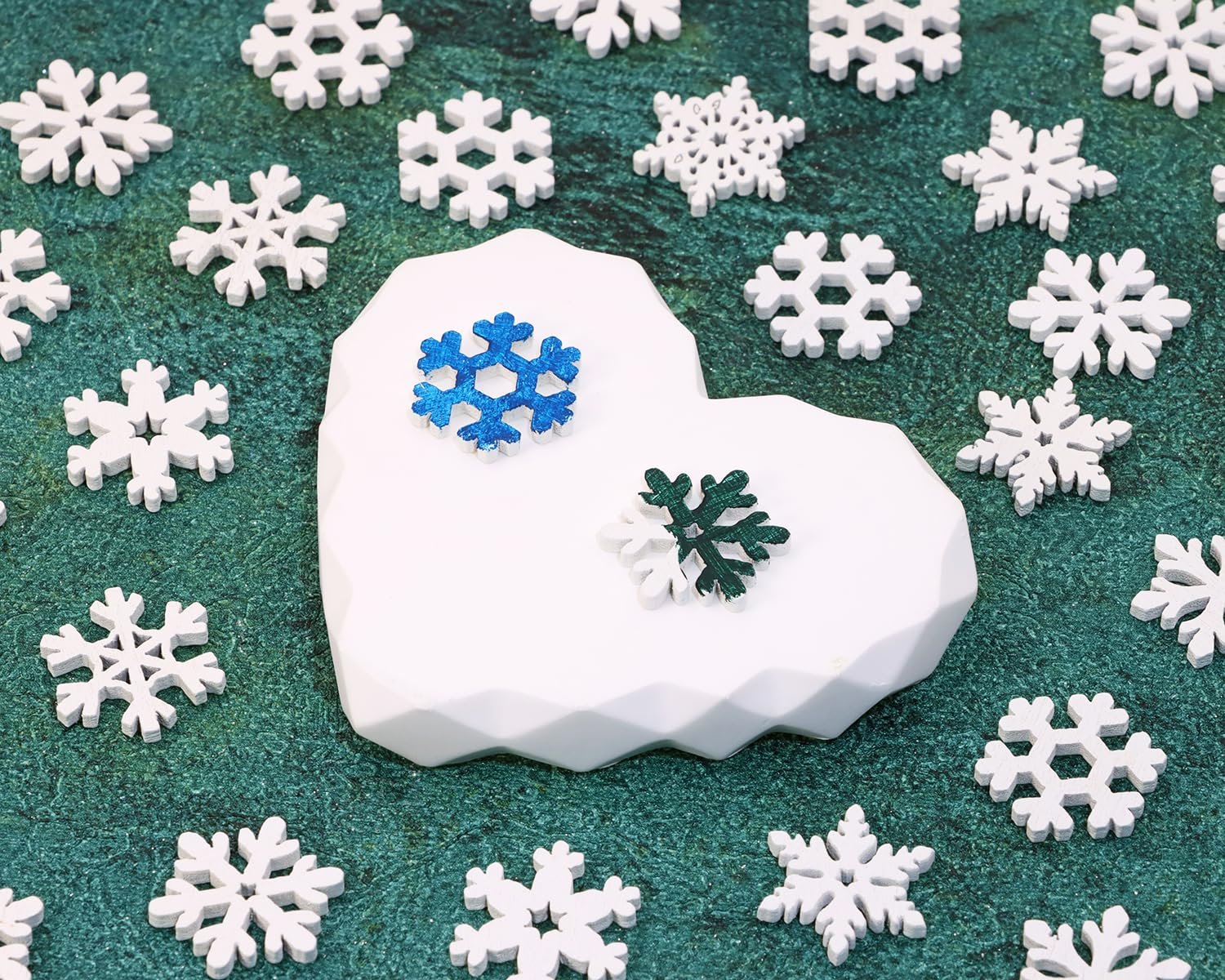 Assortment of white wooden snowflake cutouts scattered on a green textured surface, with a white geometric heart-shaped display holding two colorful snowflakes.