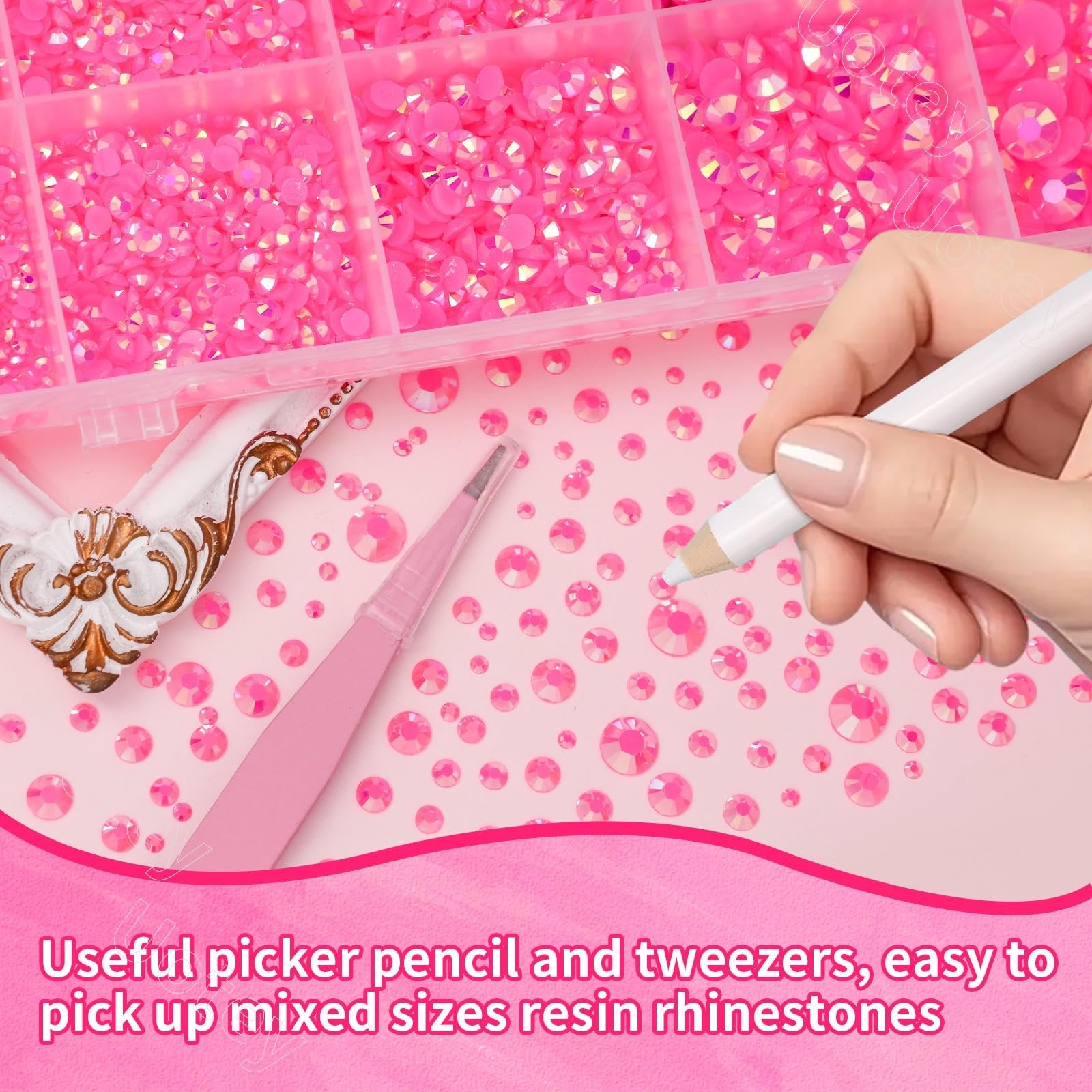 Close-up of a hand using a white pencil to pick up pink resin rhinestones from a pink surface, with tweezers and a divided container of rhinestones visible.