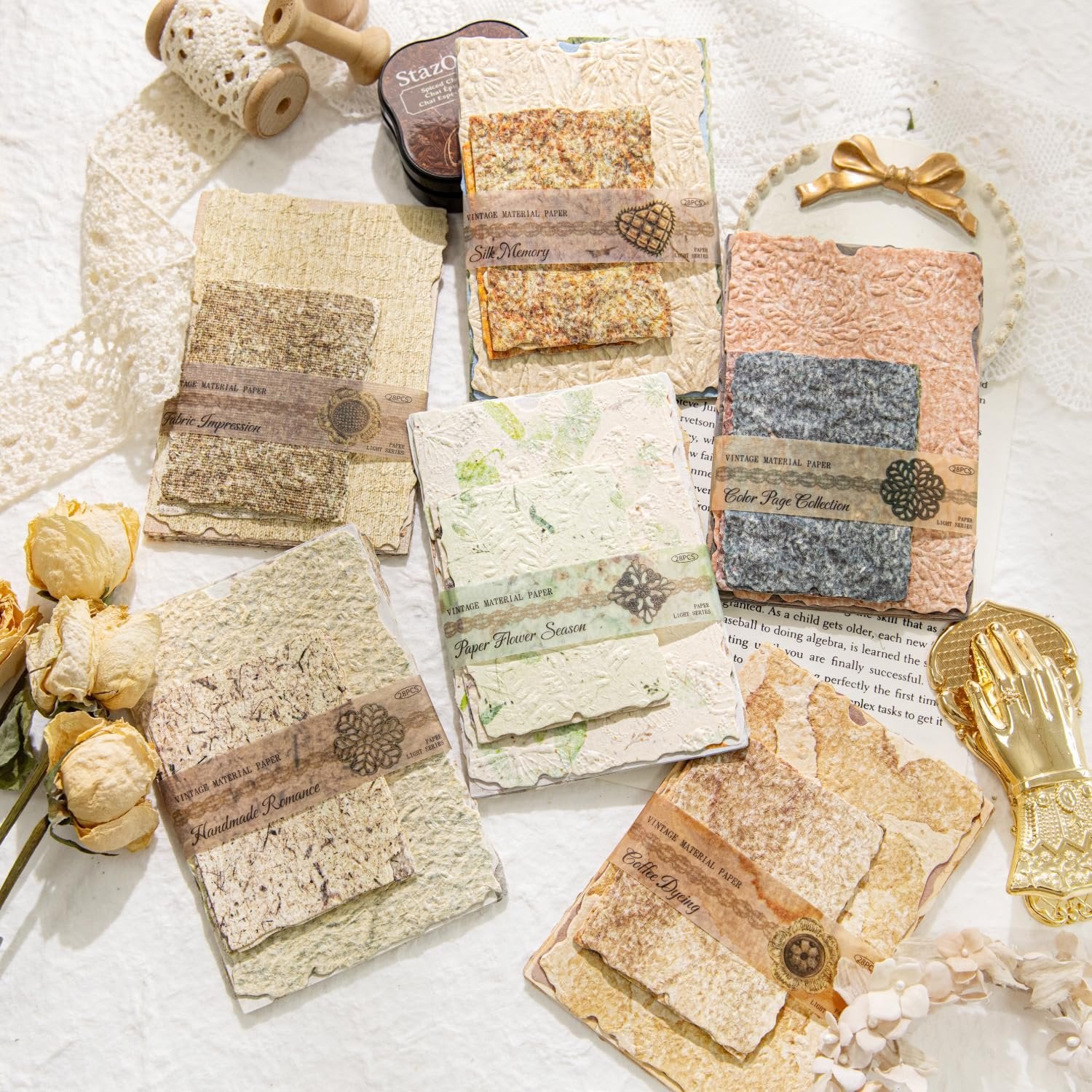 Assorted textured scrapbook paper sheets with decorative bands, arranged on a white lace cloth with vintage craft supplies.