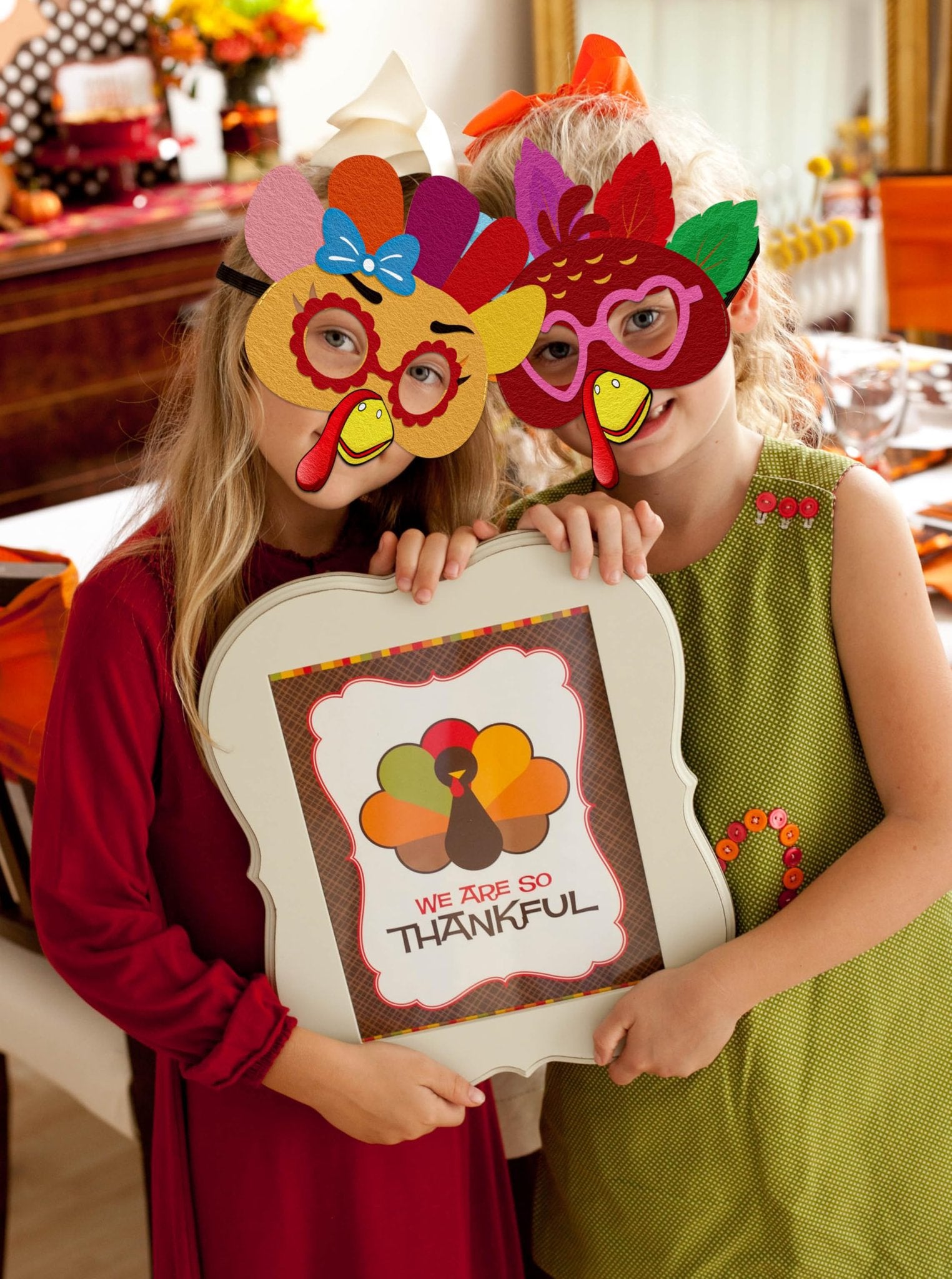 Two girls wearing DIY felt turkey masks hold a sign that says 'We Are So Thankful' at a Thanksgiving party.