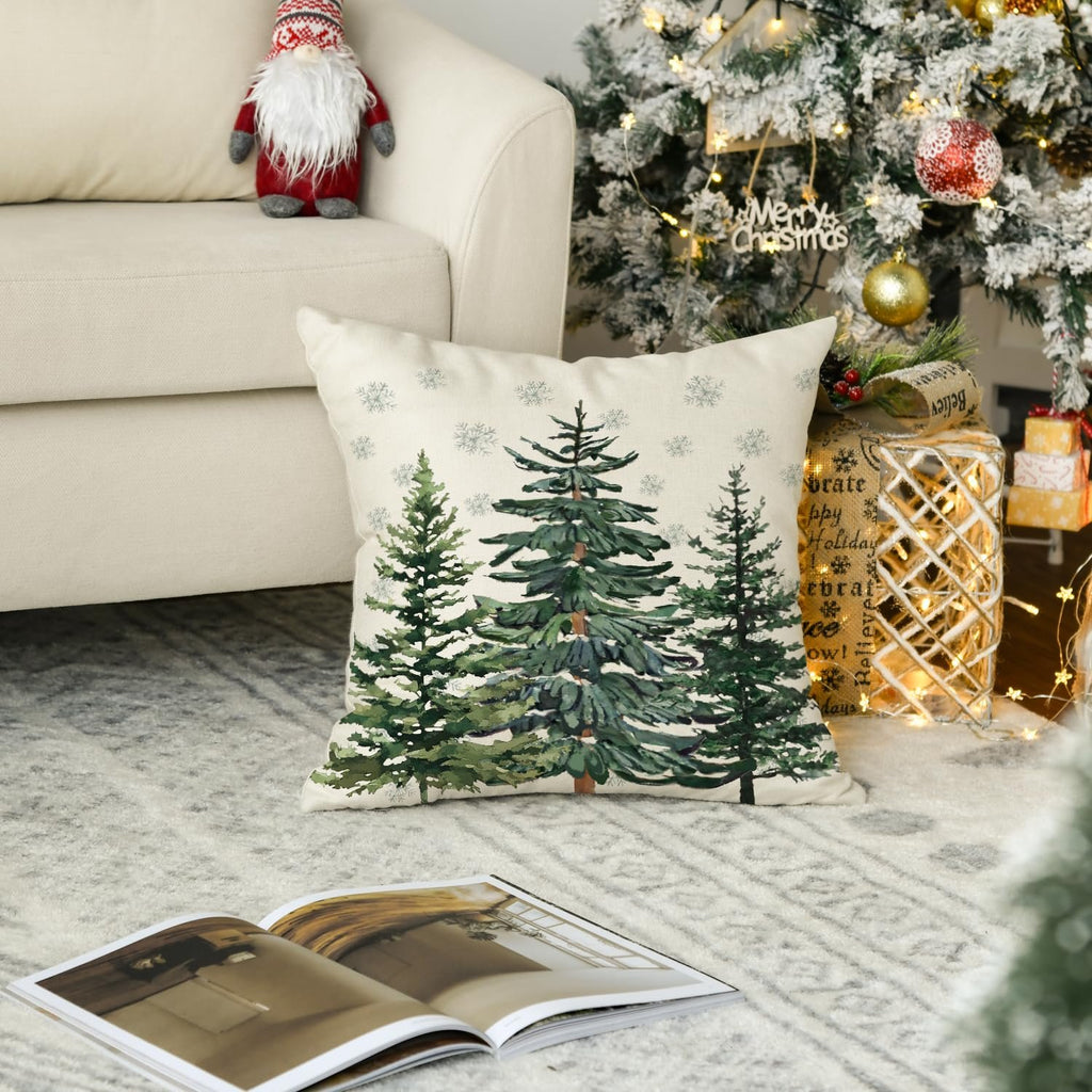 Christmas throw pillow cover with watercolor pine trees and snowflakes on a couch next to a decorated Christmas tree.
