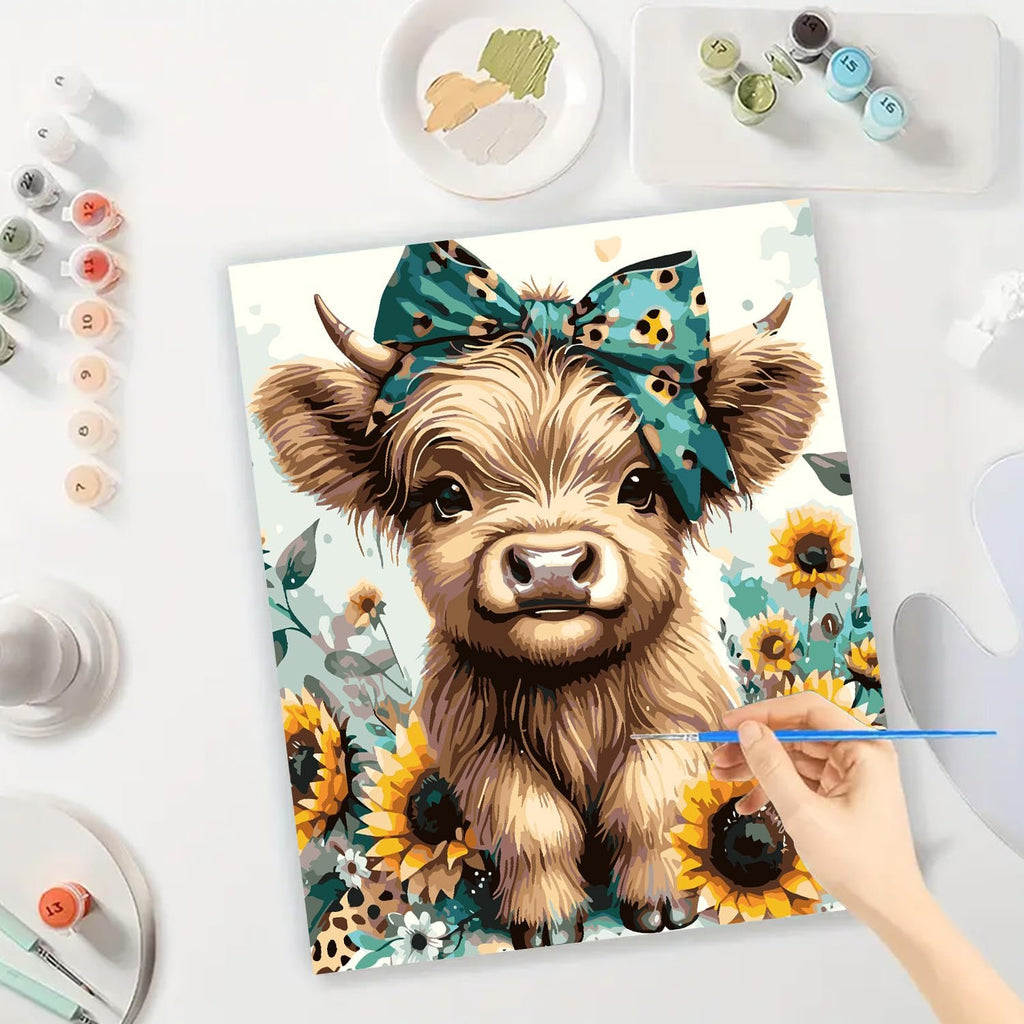 Paint by numbers kit showing a fluffy cow with a teal bow and sunflowers, with a hand holding a paintbrush.