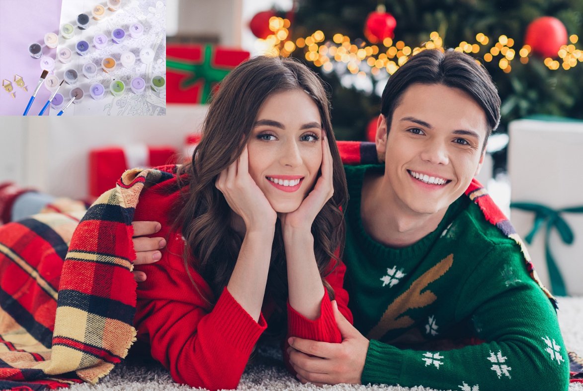 Couple in Christmas sweaters under a blanket with paint supplies and Christmas tree in background