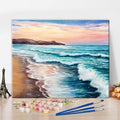 Paint by numbers kit with colorful acrylic paints, brushes, and hardware laid out on a wooden surface with a finished beach landscape painting in the background.