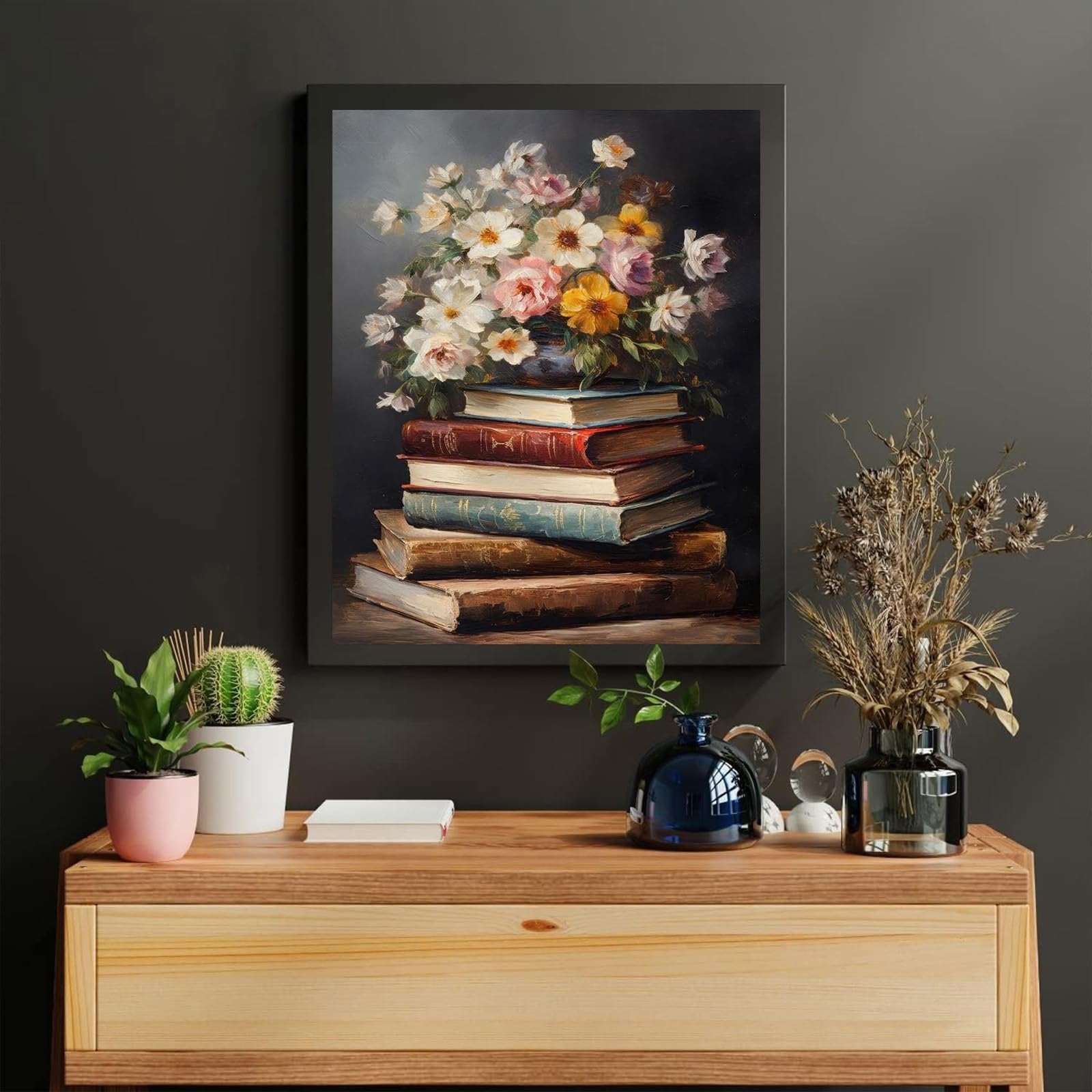 Framed painting of a stack of antique books with a floral arrangement on top, displayed above a wooden dresser with plants and vases.
