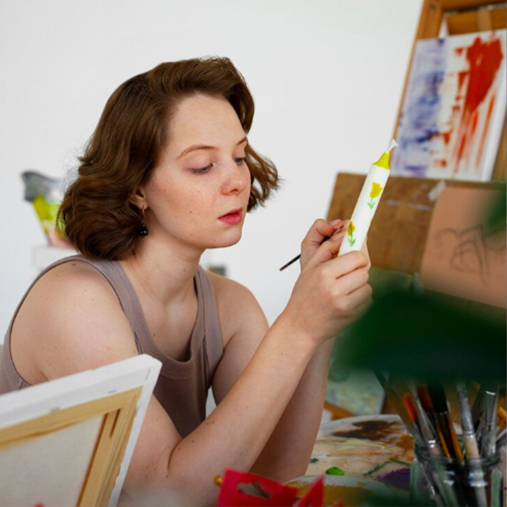 Woman painting a candle with yellow flowers, with art supplies and canvases in the background.