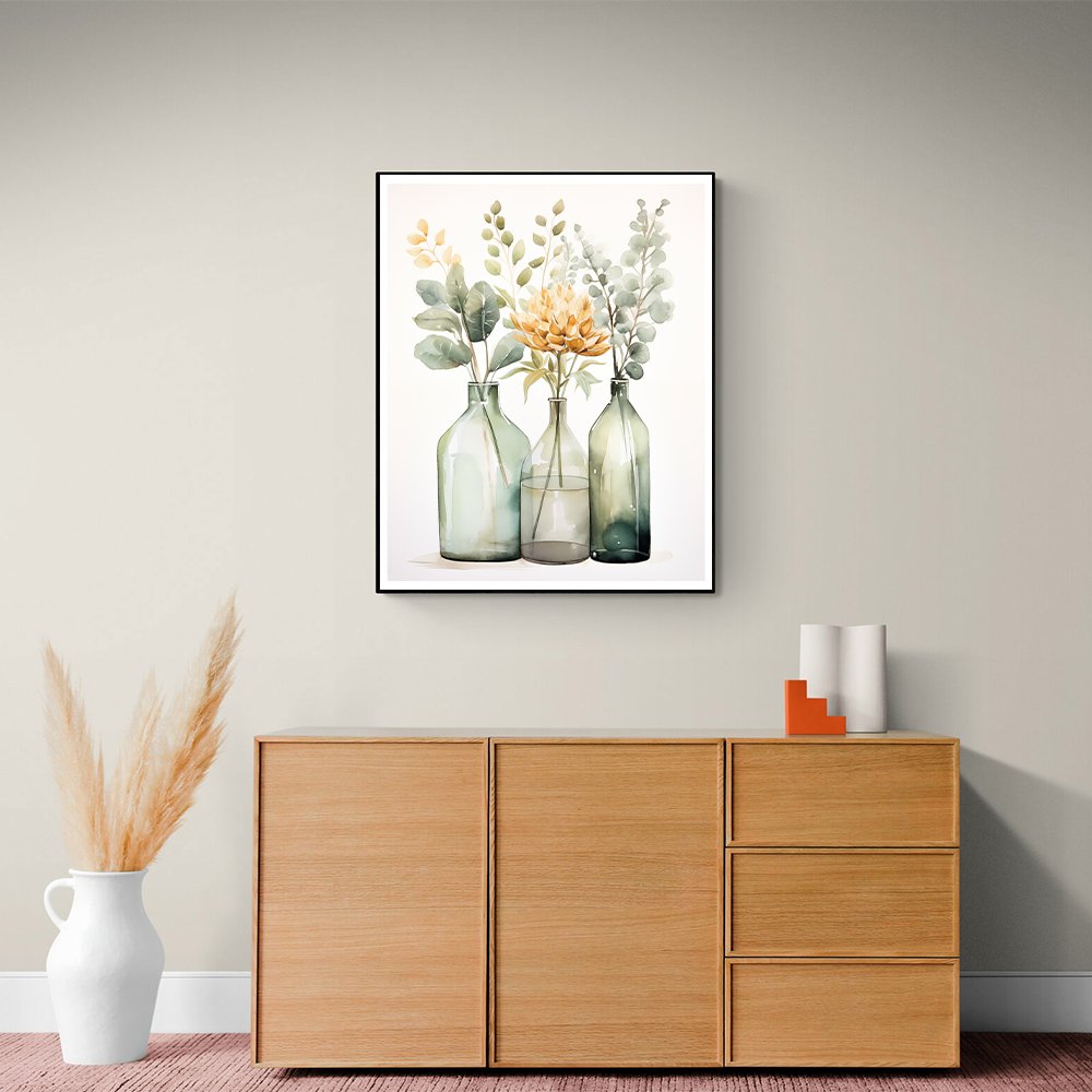 Framed watercolor painting of three glass bottles with green foliage and a yellow flower, displayed on a wall above a wooden cabinet.