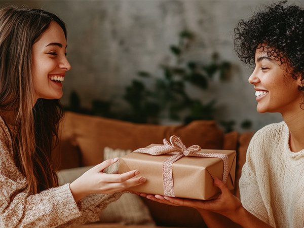 Two smiling women exchanging a wrapped gift with a pink ribbon