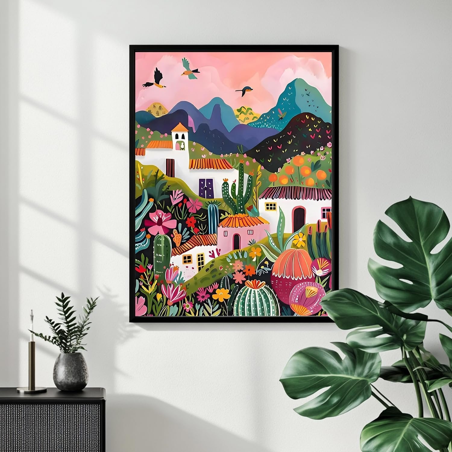 Framed colorful desert village painting hanging on a white wall, with plants in foreground.