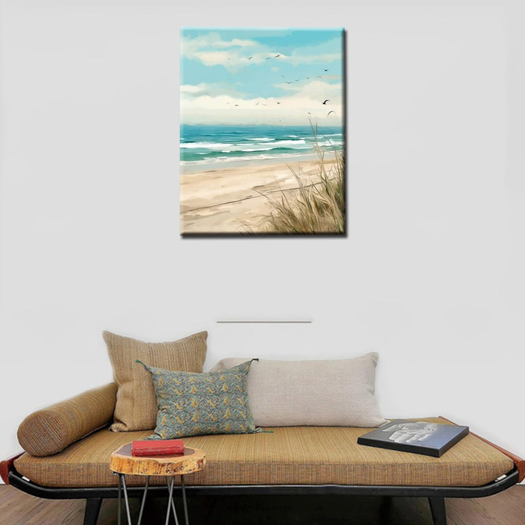 Seaside beach painting with waves, sand, and seagulls in the sky, displayed on a wall above a couch.