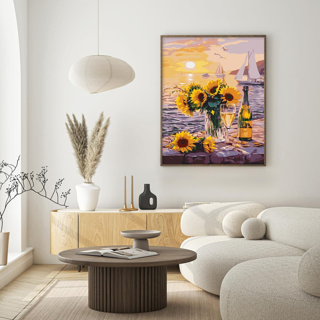 Framed paint by numbers artwork of sunflowers and champagne on a table by the sea at sunset, displayed in a modern living room.