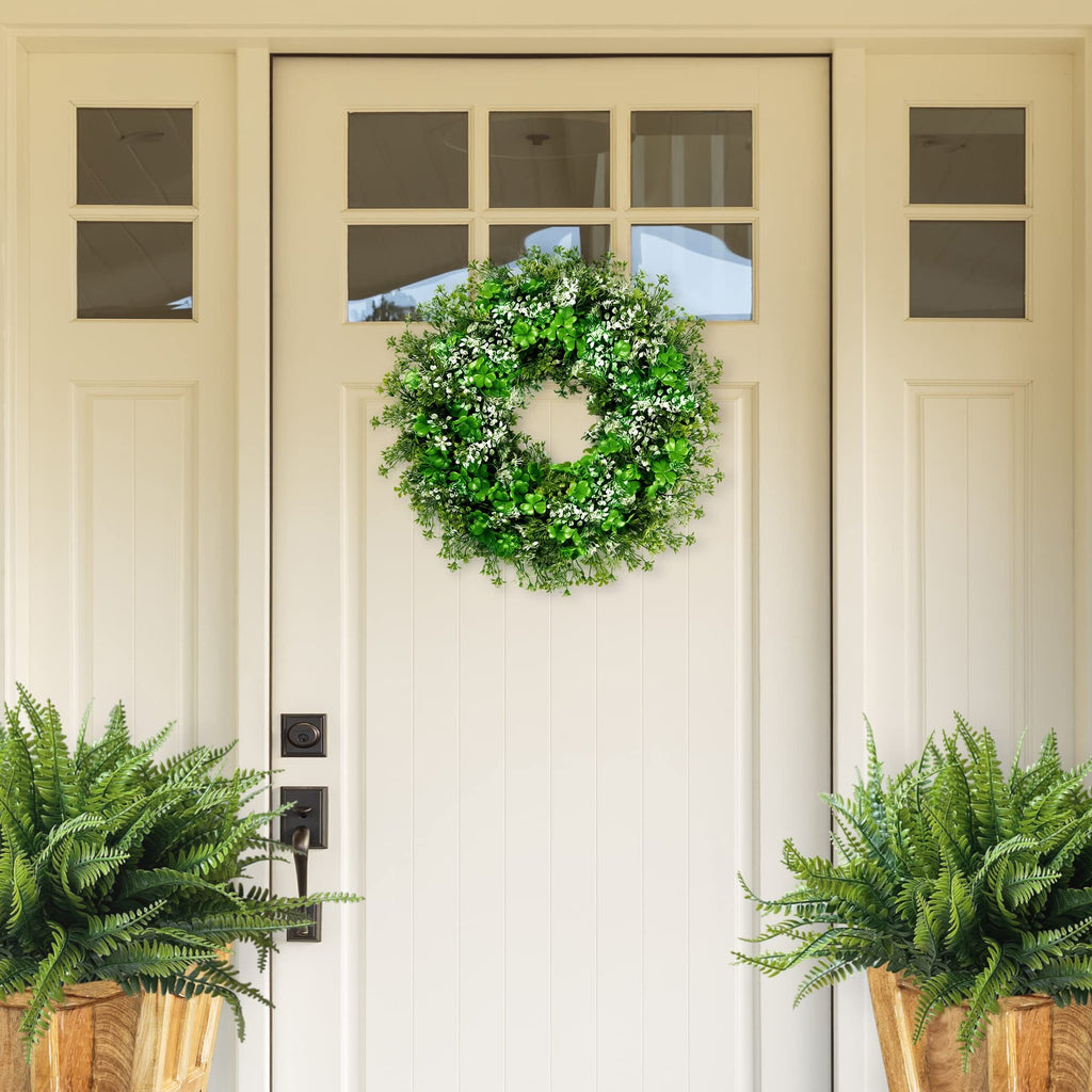 Green shamrock and flower wreath with white accents hanging on a white door, flanked by potted ferns.