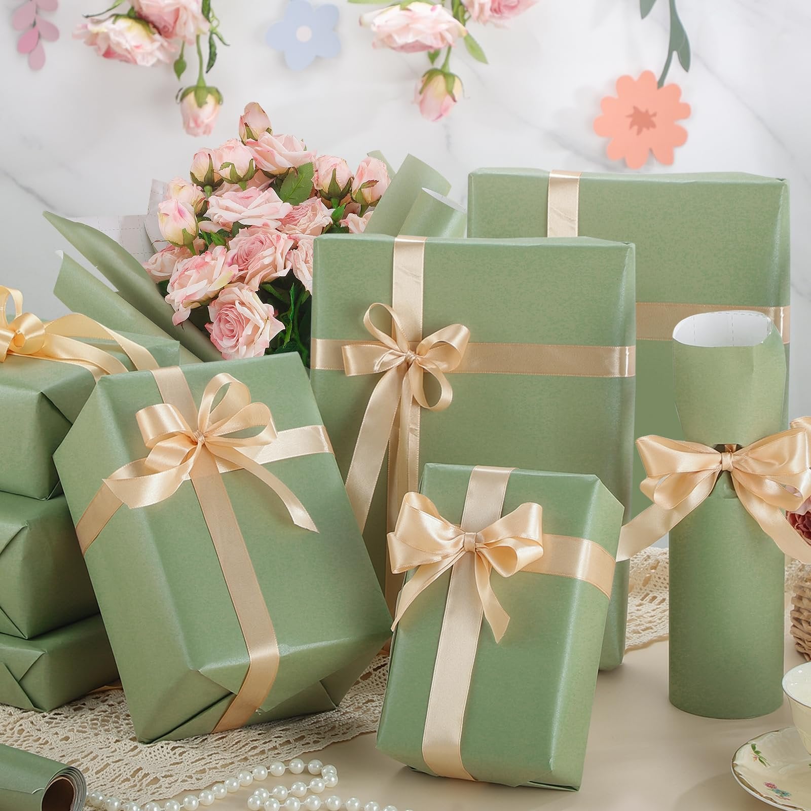 Several gift-wrapped boxes in sage green with champagne ribbon bows, arranged with a bouquet of pink roses and decorative paper flowers.