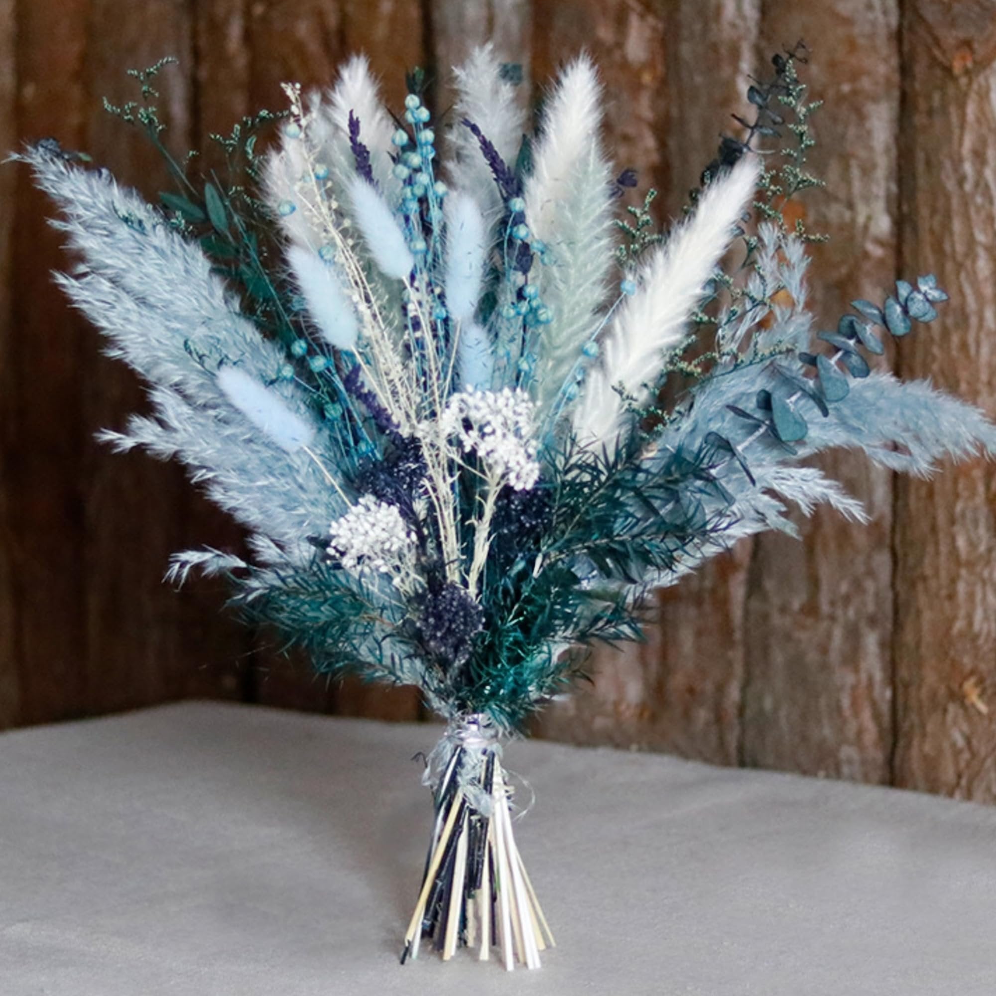 Dusty blue dried pampas grass bouquet with white grass, reeds, eucalyptus, bunny tail, money tree and dried lavender against a wooden background.