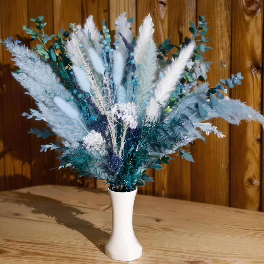 Dusty blue dried pampas grass bouquet with white grass, reeds, eucalyptus, bunny tail, and lavender in a white vase on a wooden table.