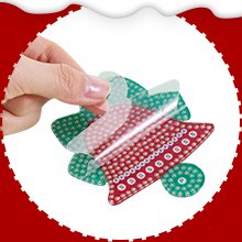Hand peeling protective film off a diamond art Christmas keychain kit shaped like a bow, with red and green sections covered in rhinestones.