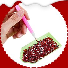 Hand holding pink tool to pick up red and white rhinestones from a green tray for diamond art crafting