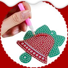 Hand using pink tool to apply diamond to red bell-shaped Christmas ornament, green bow behind