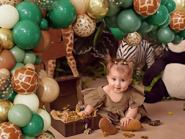 Baby girl in safari-themed outfit sits by a treasure chest with a jungle balloon arch and stuffed animals in the background.