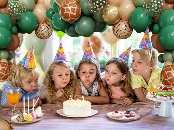 Children wearing party hats blowing out candles on a birthday cake, with a jungle safari balloon arch in the background.