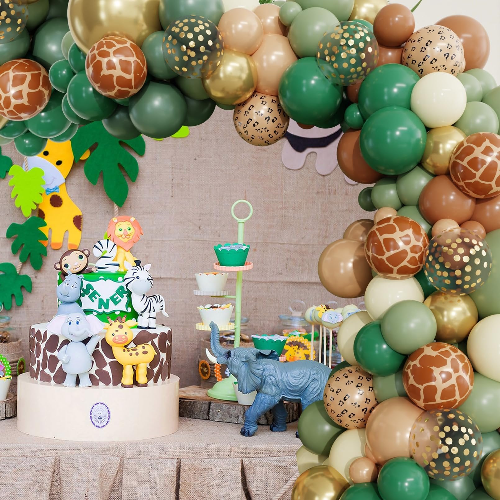 Jungle safari themed birthday cake with animal figurines and a balloon arch garland in green, brown, and gold tones.