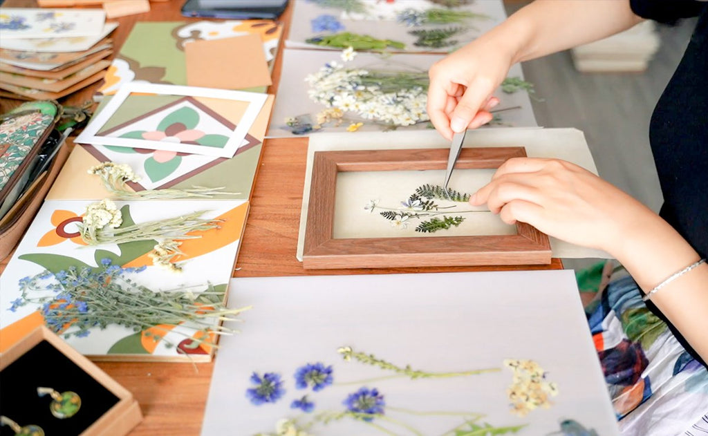 Hands using tweezers to arrange dried flowers and leaves in a wooden frame for a craft project.