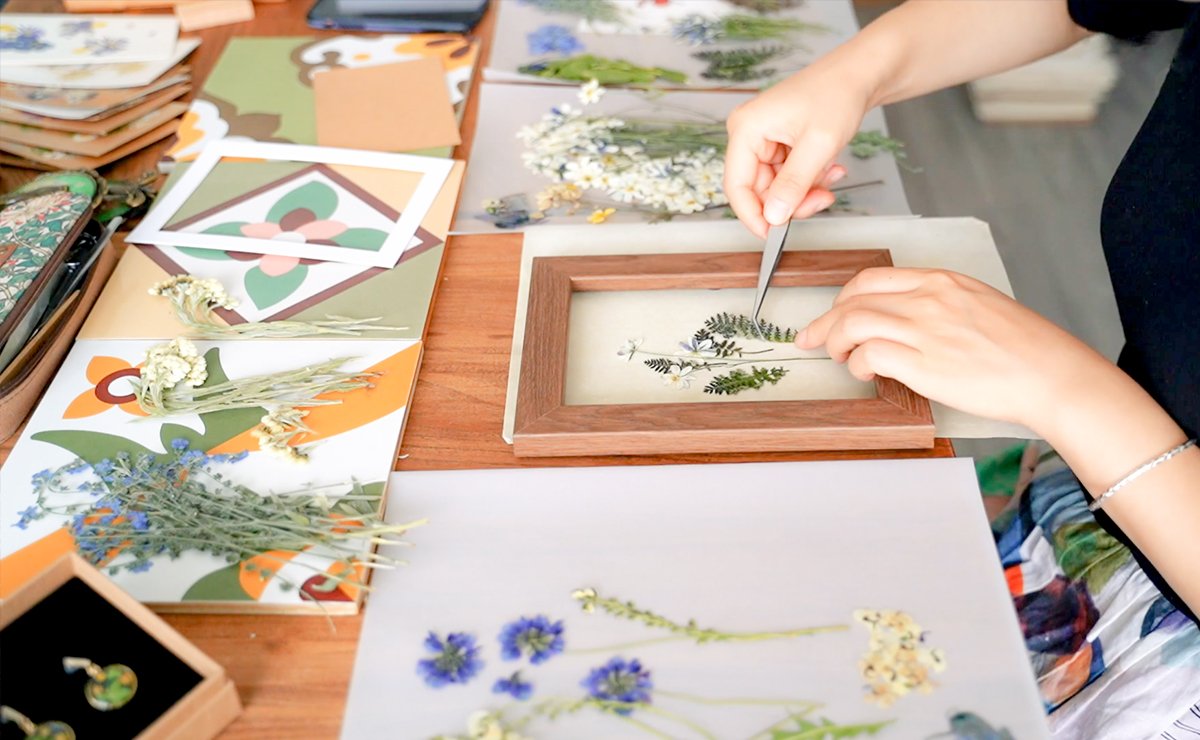 Hands using tweezers to arrange dried flowers and leaves in a wooden frame for a craft project.