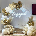 Balloon arch with white, gold, and sand-colored balloons, featuring a neon 'Engaged' sign and dried palm leaves.