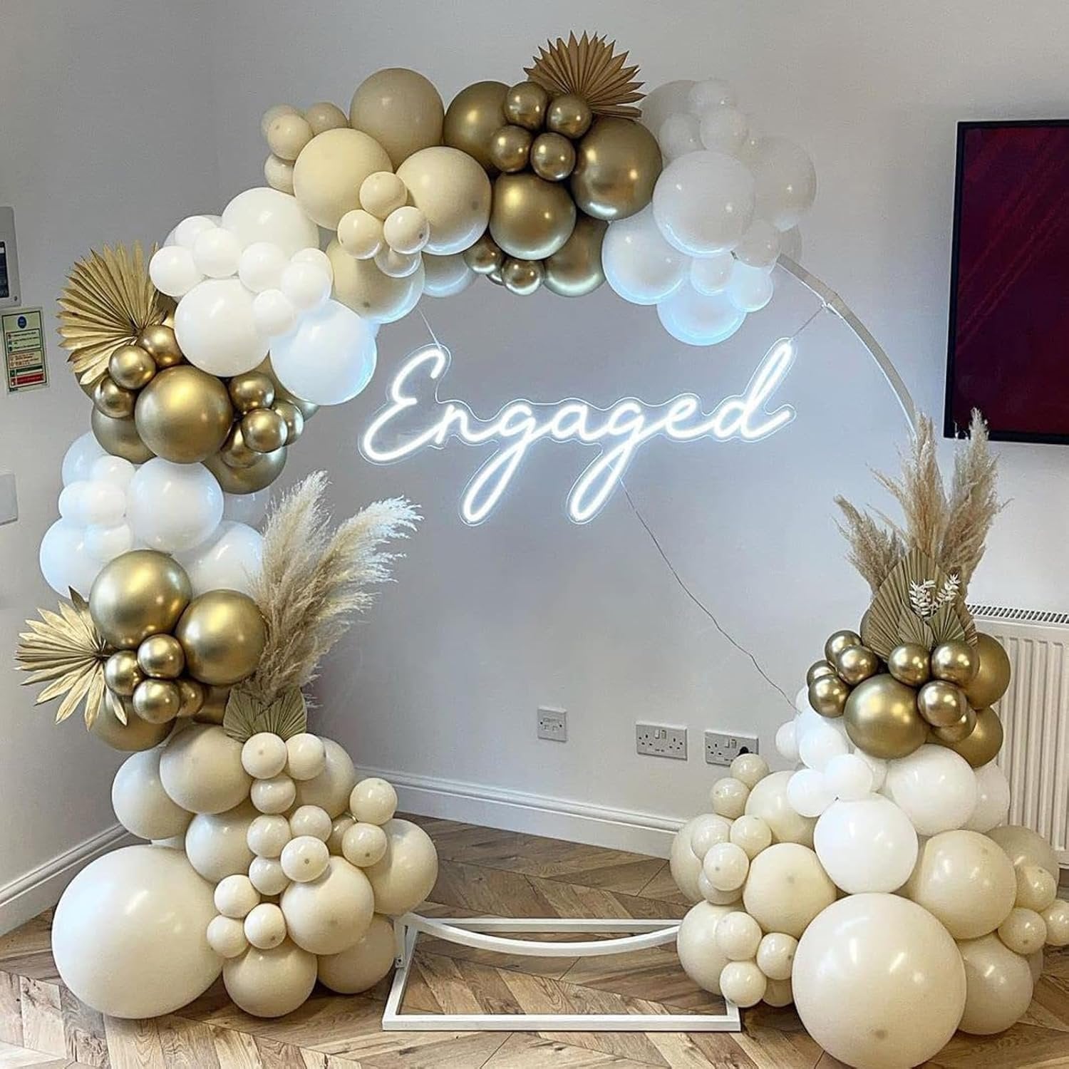 Balloon arch with white, gold, and sand-colored balloons, featuring a neon 'Engaged' sign and dried palm leaves.