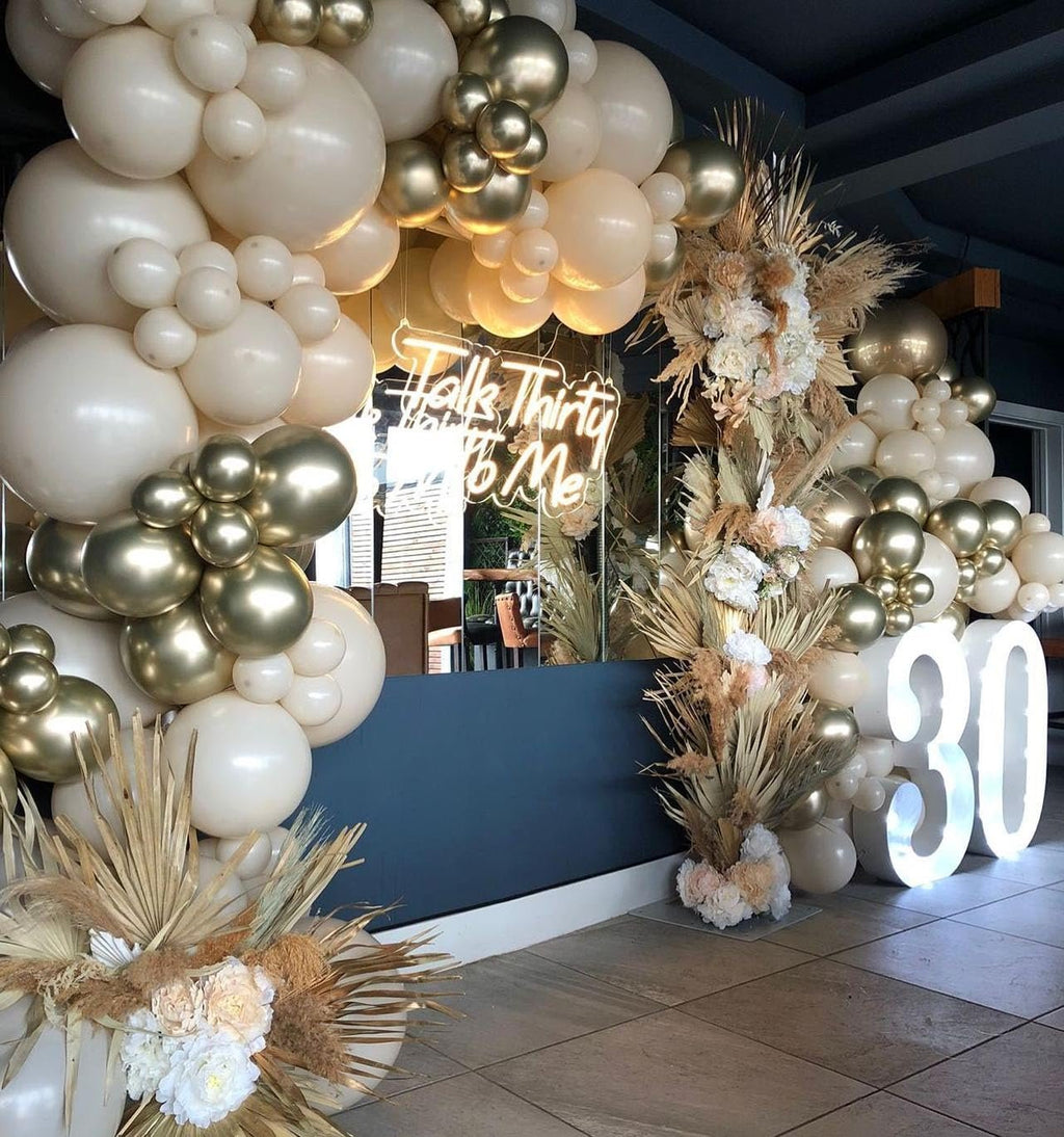 Balloon arch in sand white and gold with large illuminated '30' numbers and a neon sign saying 'Talk Thirty To Me'