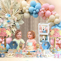 Two babies at a gender reveal party table with pink and blue balloons, cake, and decorations.