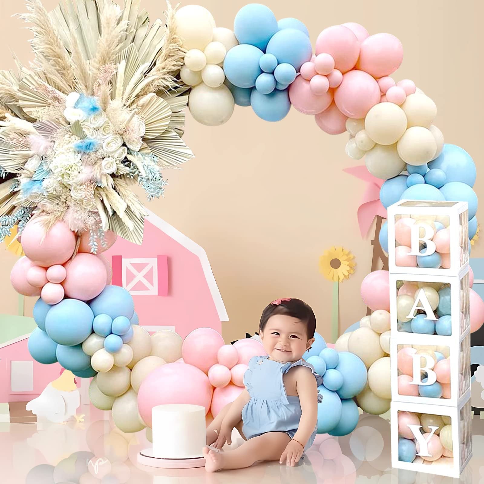 Gender reveal party setup with a baby girl sitting near a balloon arch, baby blocks spelling 'BABY', and a pink barn backdrop.