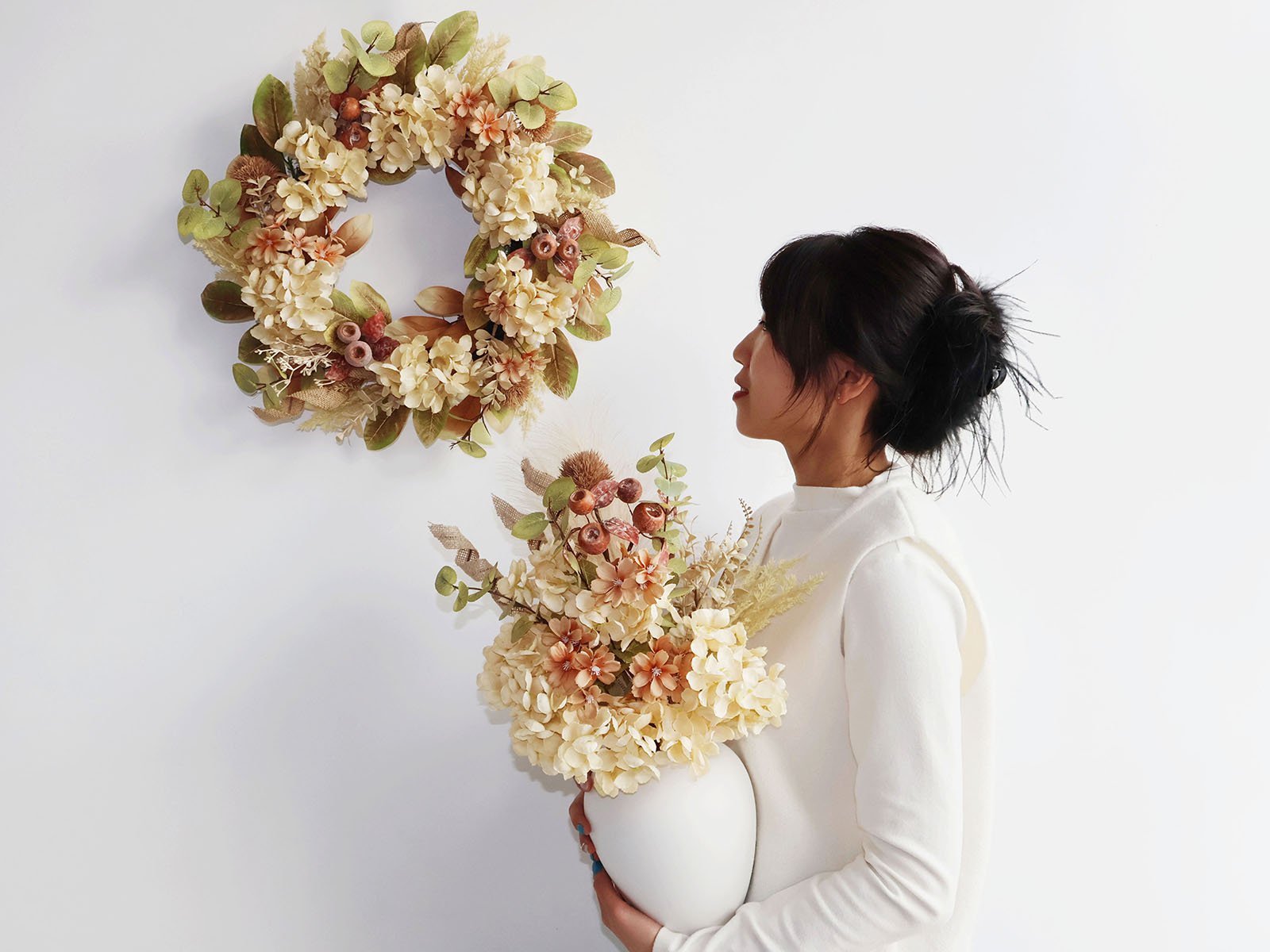 Woman holding a vase of fall flowers next to a hanging fall wreath