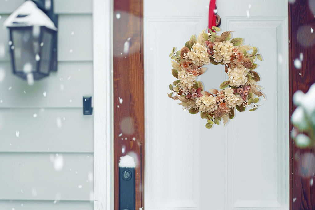 Fall wreath with faux hydrangea, pampas grass, and burlap hanging on a white door during snowfall.