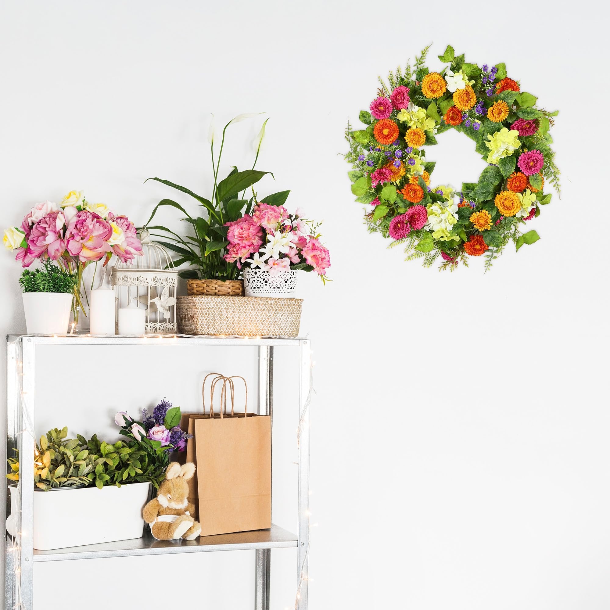 Artificial daisy wreath with green leaves hanging on a white wall above a shelf with flowers and decor