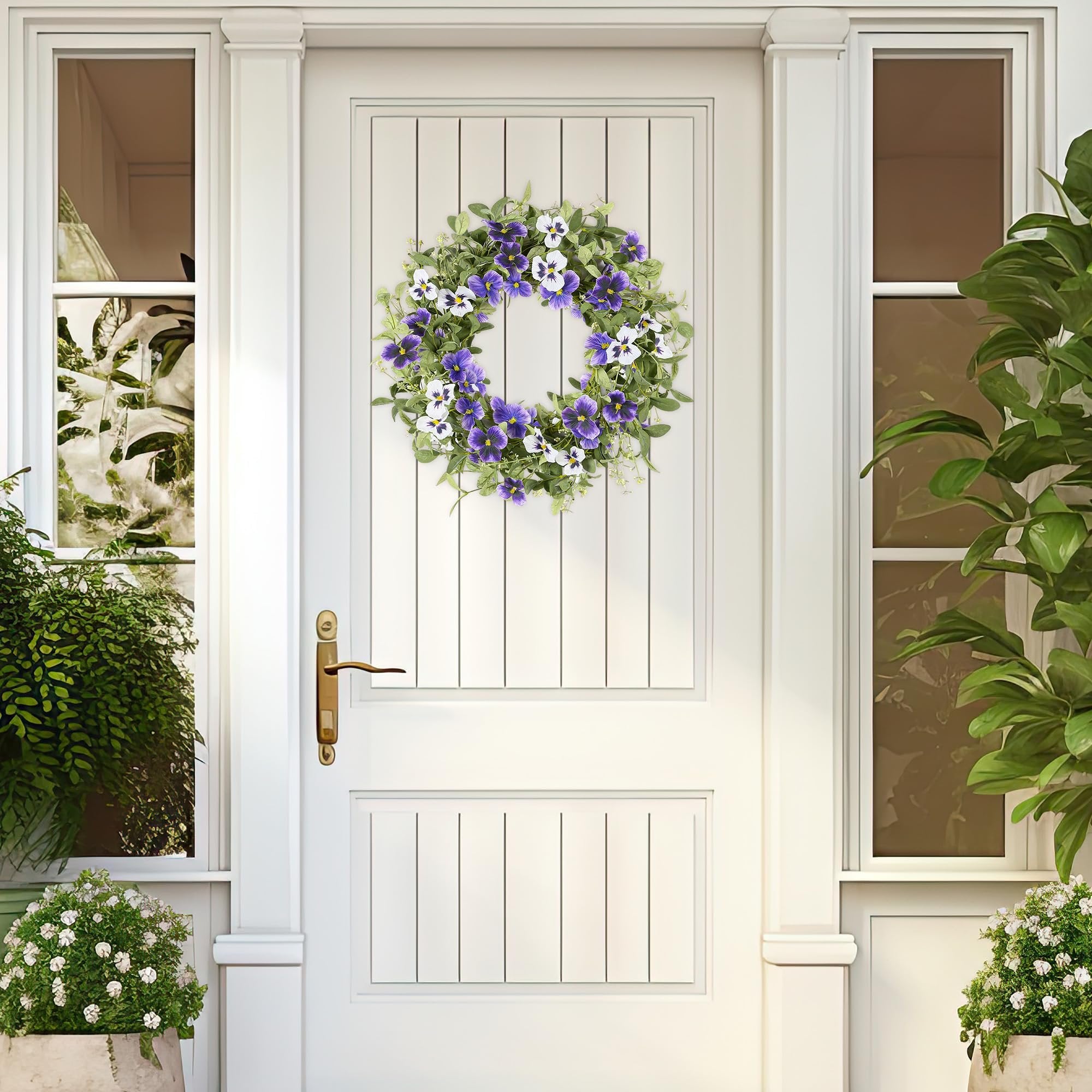 Artificial floral wreath with purple and white pansies and green leaves hanging on a white paneled door.