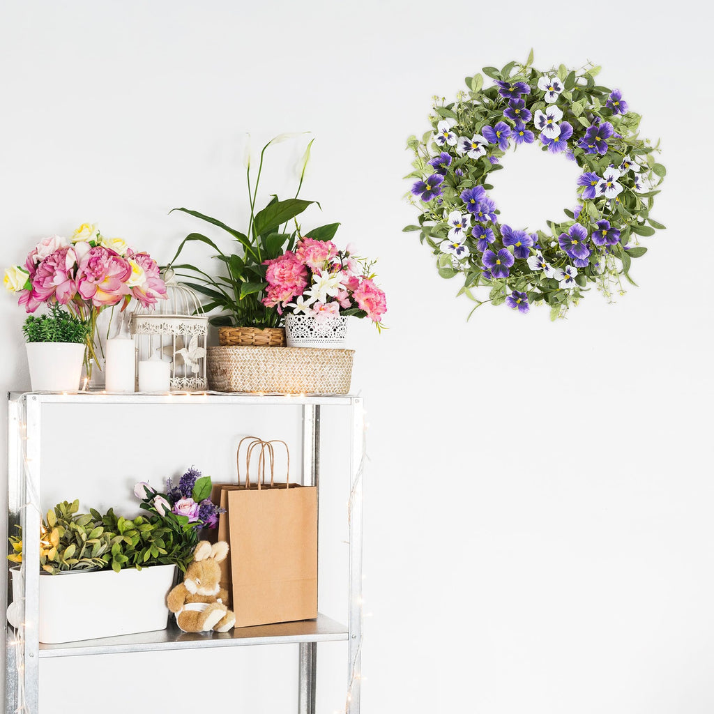 Artificial floral wreath with purple pansies and green leaves hanging on a white wall above a shelf with decorative items.