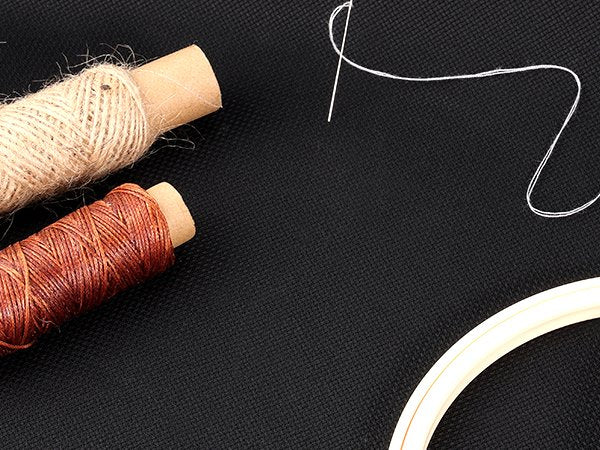 Two spools of thread and a needle with white thread on a black textured surface, with a portion of an embroidery hoop visible.
