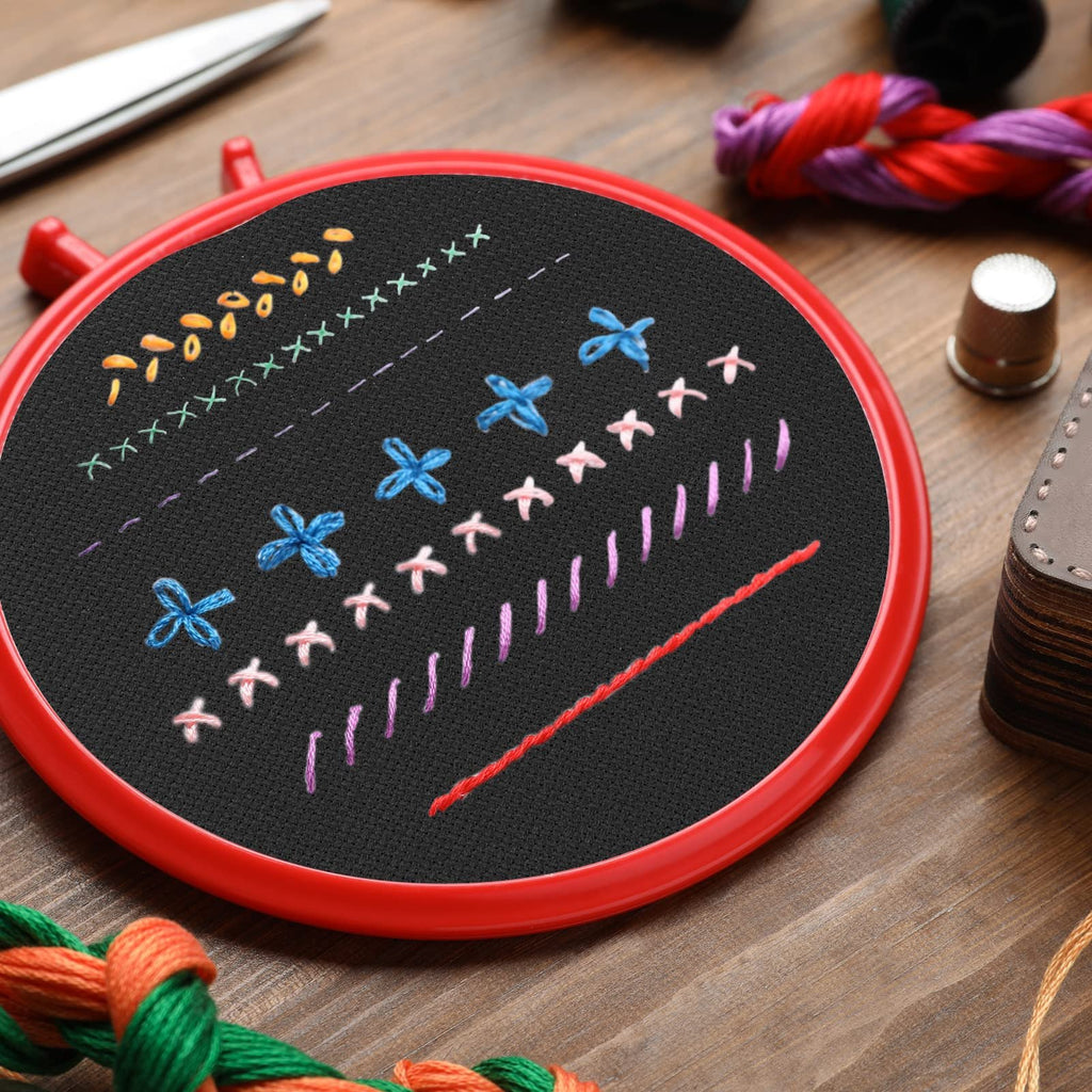 Embroidery hoop with black Aida cloth showing various stitched patterns and embroidery floss, scissors, and thimble on wooden surface.
