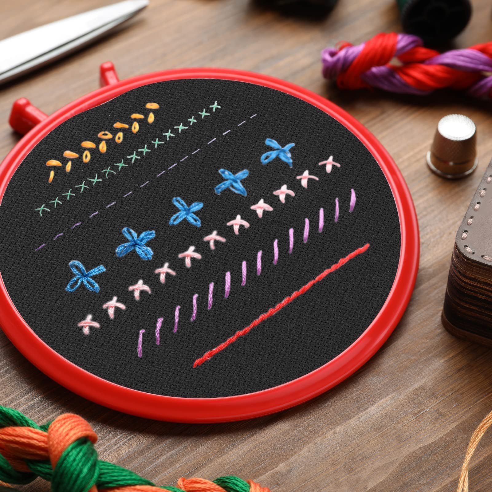 Embroidery hoop with black Aida cloth showing various stitched patterns and embroidery floss, scissors, and thimble on wooden surface.