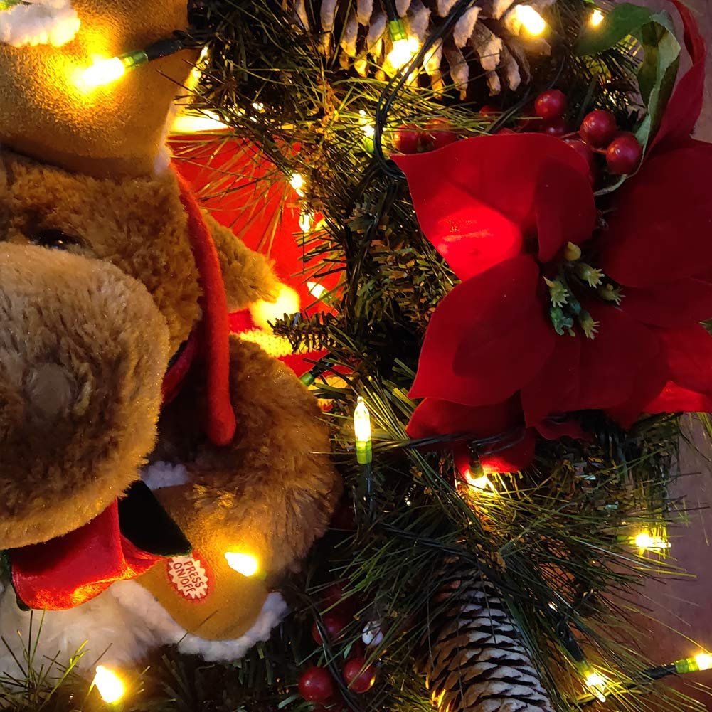 Close-up of a plush reindeer toy with a red scarf, surrounded by warm white LED string lights, pinecones, and red poinsettias.