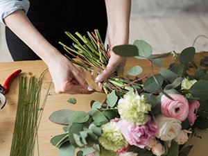 Hands arranging a bouquet of flowers and greenery with floral stem wire on a wooden table.
