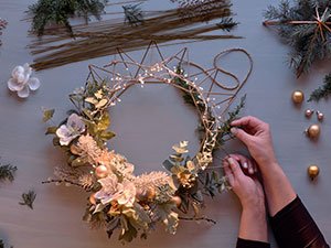 Hands crafting a star-shaped wreath with fairy lights, faux flowers, and greenery on a flat surface.