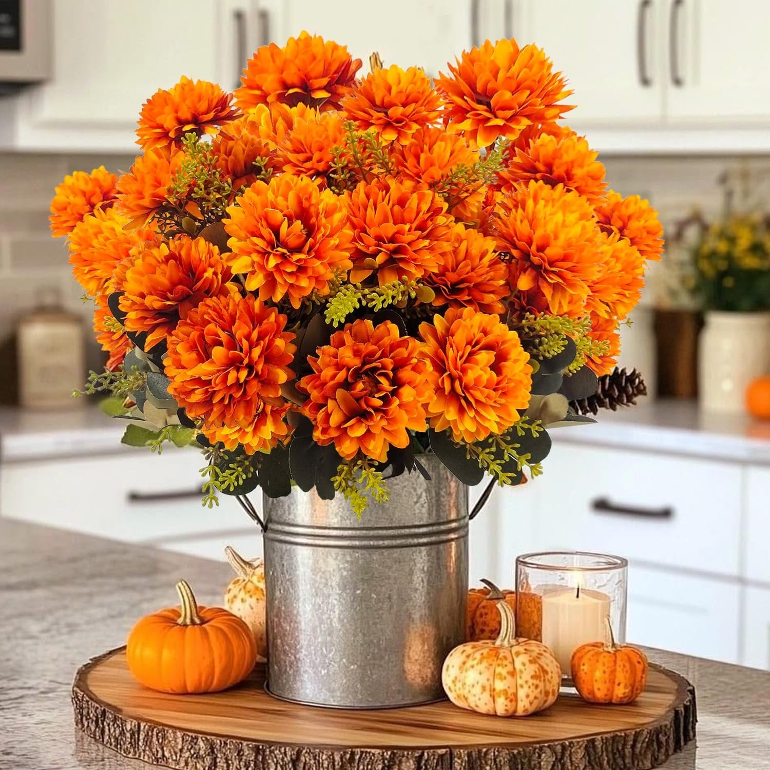 Artificial orange mum bouquet in a galvanized bucket with pumpkins and a candle on a wood slice