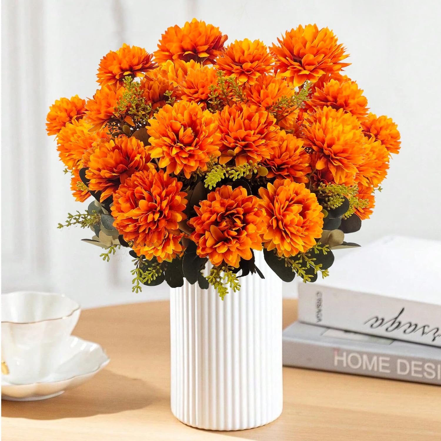 Orange artificial mum flowers in a white vase with greenery, next to a teacup and books.