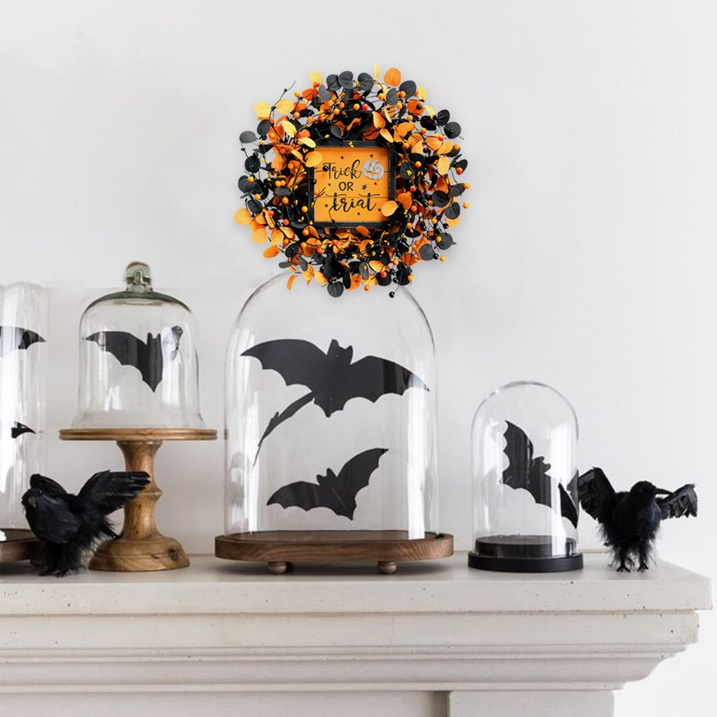Halloween wreath with orange and black leaves and berries, featuring a 'Trick or Treat' sign in the center, displayed above glass domes with bat silhouettes.