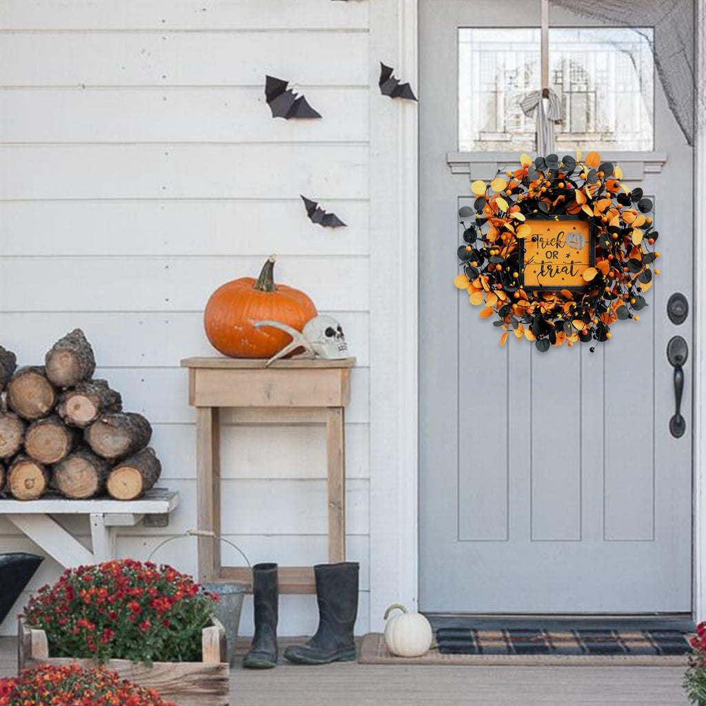 Halloween wreath with orange and black eucalyptus leaves and berries hanging on a gray door, with a pumpkin and skull on a table nearby.